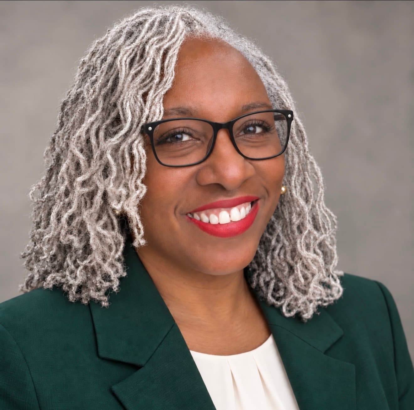 Portrait of a smiling woman with silver-gray locs, black glasses, and a green blazer